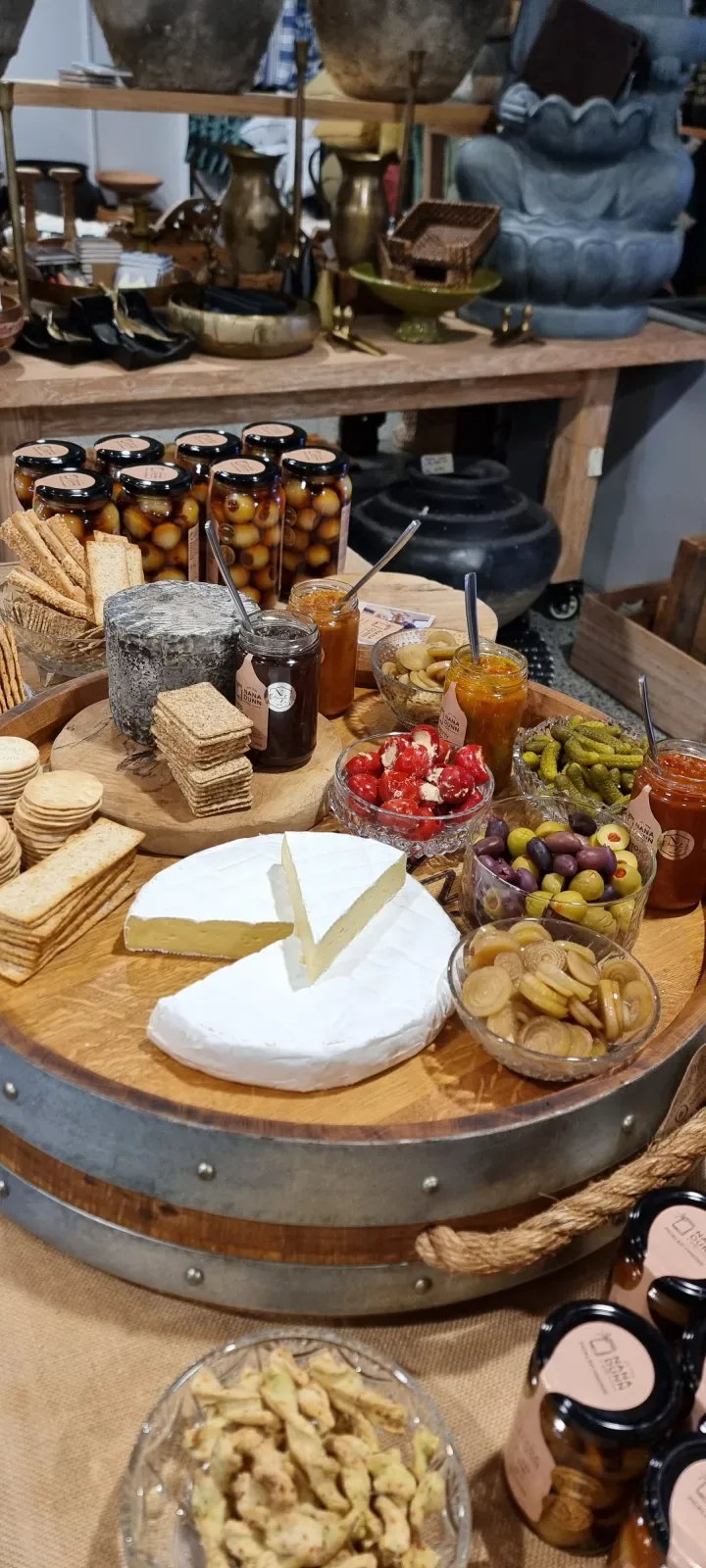A platter on a barrel table at Cote Bleu Home, with cheese, crackers, olives and jars of Nana Dunn + Co pickles and relishes