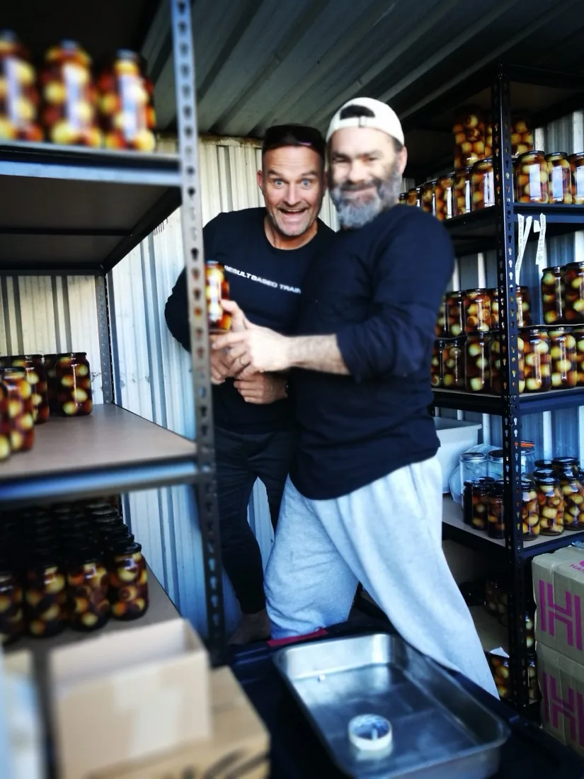 Scott and Wayne holding a jar among shelves of Nana Dunn jars in a storeroom shed